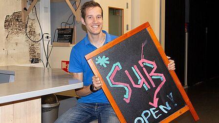 Micheal Laan aan de bar in De Schoof met een krijtbord in de handen met 'SUB open' erop geschreven.